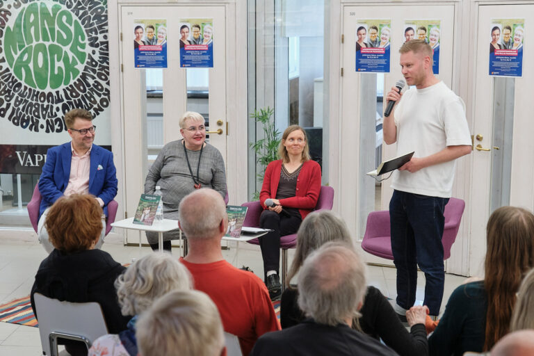 Paneelikeskustelijat ja moderaattori hymyilevät yleisön edessä.